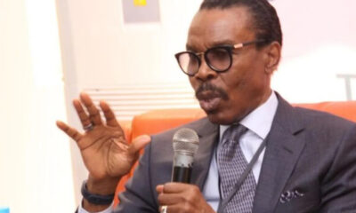 Man in a dark suit and glasses speaks into a handheld microphone, gesturing with his left hand.