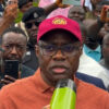 Man in an orange polo shirt and red cap speaks into a microphone while a crowd records him with smartphones behind.