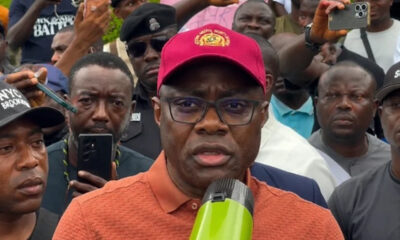 Man in an orange polo shirt and red cap speaks into a microphone while a crowd records him with smartphones behind.