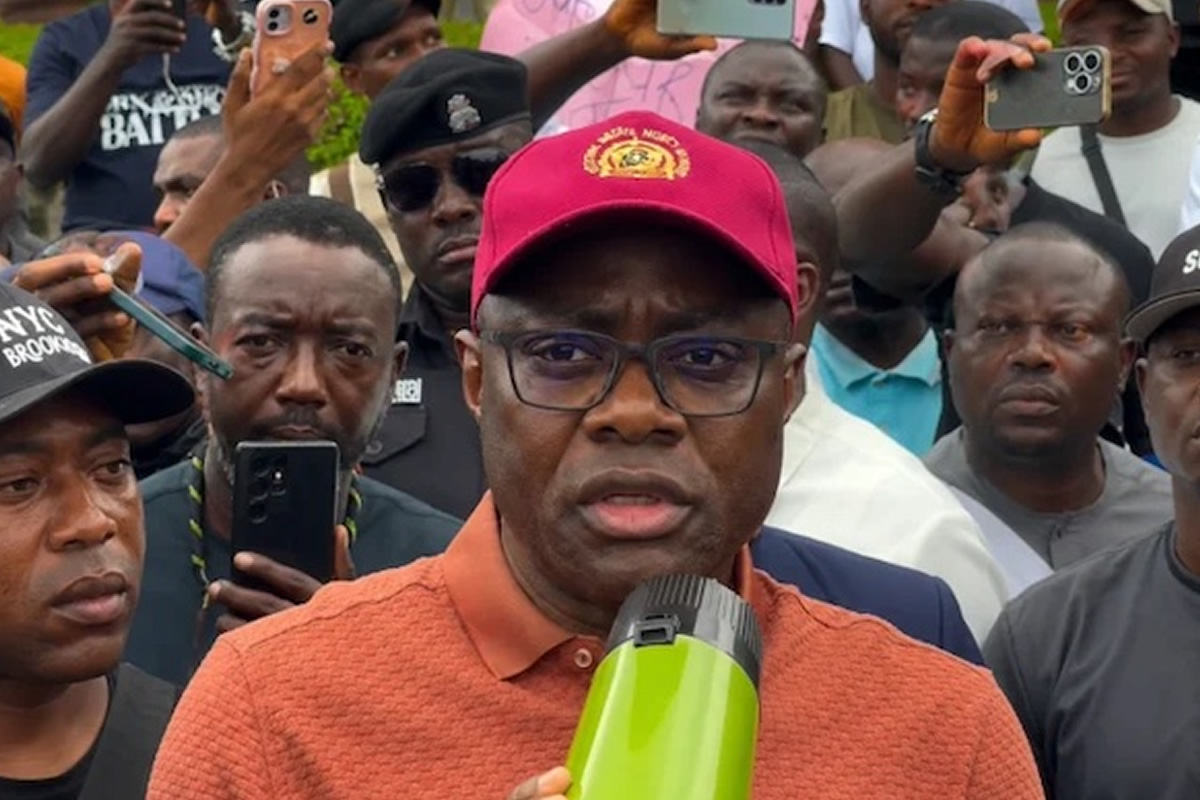 Man in an orange polo shirt and red cap speaks into a microphone while a crowd records him with smartphones behind.