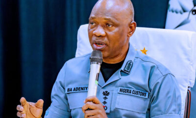 Nigeria Customs officer in uniform speaking into a handheld microphone at a formal event, seated at a table with a white chair cover behind him.