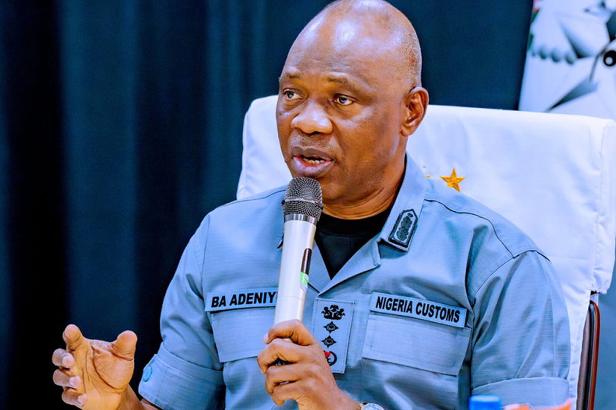 Nigeria Customs officer in uniform speaking into a handheld microphone at a formal event, seated at a table with a white chair cover behind him.