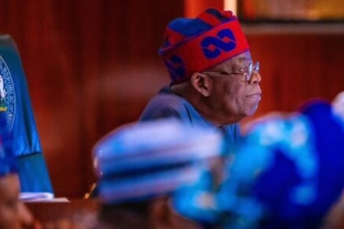 Side profile of an older man wearing a red and blue traditional cap and glasses, seated in a meeting room.