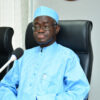 Man in light-blue traditional outfit and cap seated in a high-backed chair, speaking into a microphone on the desk ahead of him.