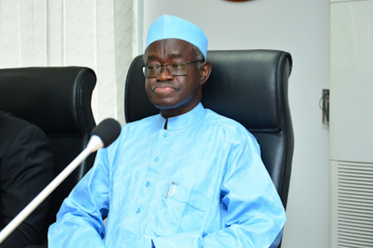 Man in light-blue traditional outfit and cap seated in a high-backed chair, speaking into a microphone on the desk ahead of him.