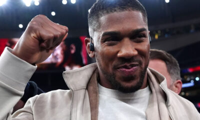 Man on a sports stage raises a clenched fist in celebration, wearing a light beige jacket and white shirt with an in-ear monitor nearby.