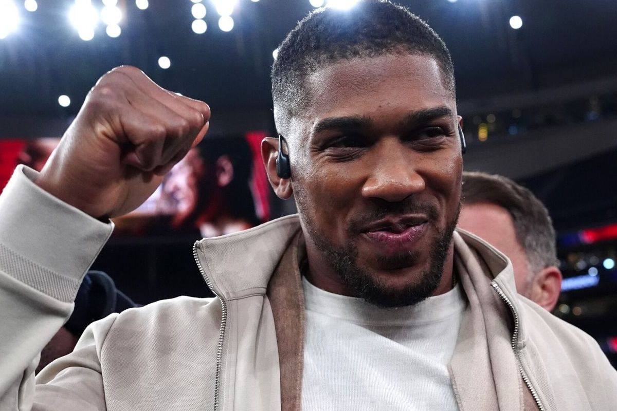 Man on a sports stage raises a clenched fist in celebration, wearing a light beige jacket and white shirt with an in-ear monitor nearby.