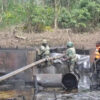 Workers in camouflage and safety vests contain an oil spill in a muddy, wooded area with large metal pipes and a wooden beam.