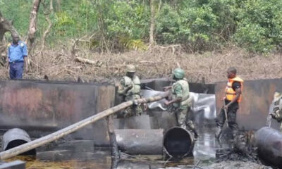 Workers in camouflage and safety vests contain an oil spill in a muddy, wooded area with large metal pipes and a wooden beam.