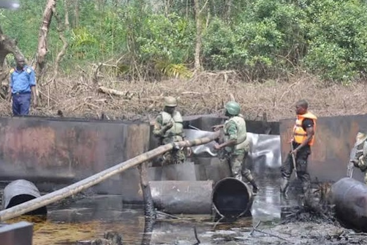 Workers in camouflage and safety vests contain an oil spill in a muddy, wooded area with large metal pipes and a wooden beam.