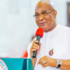 Man in white traditional attire speaks into a red microphone at a podium with a government seal visible on the front.