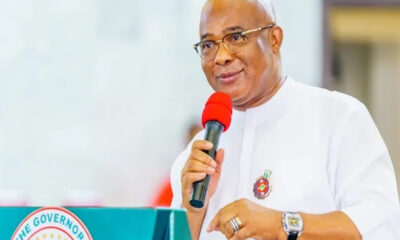 Man in white traditional attire speaks into a red microphone at a podium with a government seal visible on the front.