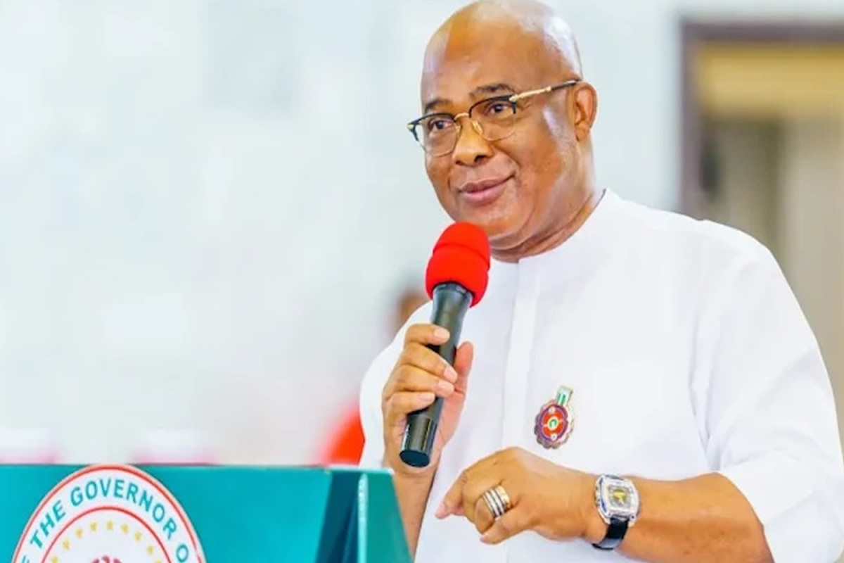 Man in white traditional attire speaks into a red microphone at a podium with a government seal visible on the front.