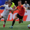 Two soccer players sprint for the ball during a match: a white-jersey defender (number 2) from one team and a red-jersey attacker (number 10) from the opposing team close behind.