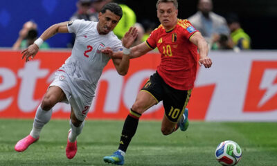 Two soccer players sprint for the ball during a match: a white-jersey defender (number 2) from one team and a red-jersey attacker (number 10) from the opposing team close behind.