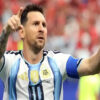 Argentinian footballer in a light blue and white jersey with a captain's armband raises both arms and points, celebrating a goal.