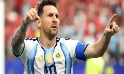 Argentinian footballer in a light blue and white jersey with a captain's armband raises both arms and points, celebrating a goal.