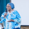 Woman wearing a blue headwrap and lace gown speaks into a microphone at a presentation, with a projector screen behind.