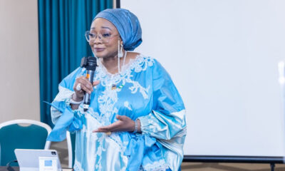 Woman wearing a blue headwrap and lace gown speaks into a microphone at a presentation, with a projector screen behind.