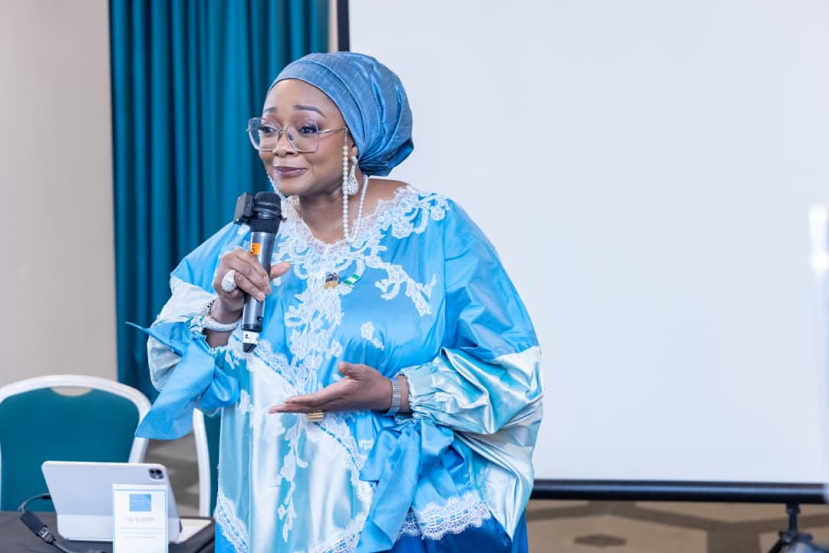 Woman wearing a blue headwrap and lace gown speaks into a microphone at a presentation, with a projector screen behind.