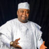 Smiling man in a white traditional agbada and patterned cap, gesturing with open hands against a dark background.] ,