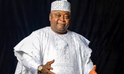 Smiling man in a white traditional agbada and patterned cap, gesturing with open hands against a dark background.] ,