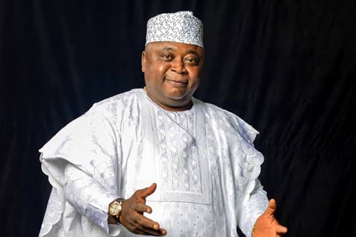 Smiling man in a white traditional agbada and patterned cap, gesturing with open hands against a dark background.] ,