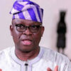 Portrait of a man wearing glasses, a white embroidered agbada, and a purple patterned fila, speaking to the camera.
