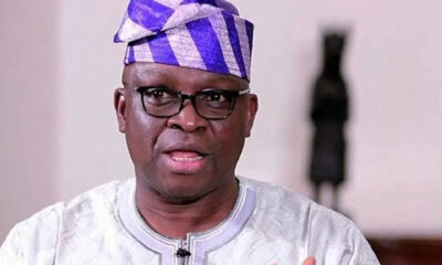 Portrait of a man wearing glasses, a white embroidered agbada, and a purple patterned fila, speaking to the camera.