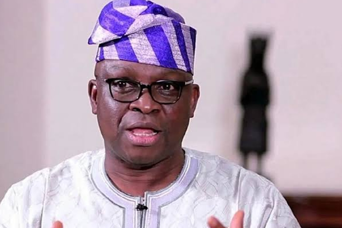 Portrait of a man wearing glasses, a white embroidered agbada, and a purple patterned fila, speaking to the camera.