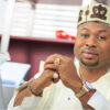 Man wearing traditional Nigerian attire and cap sits at a desk with hands clasped