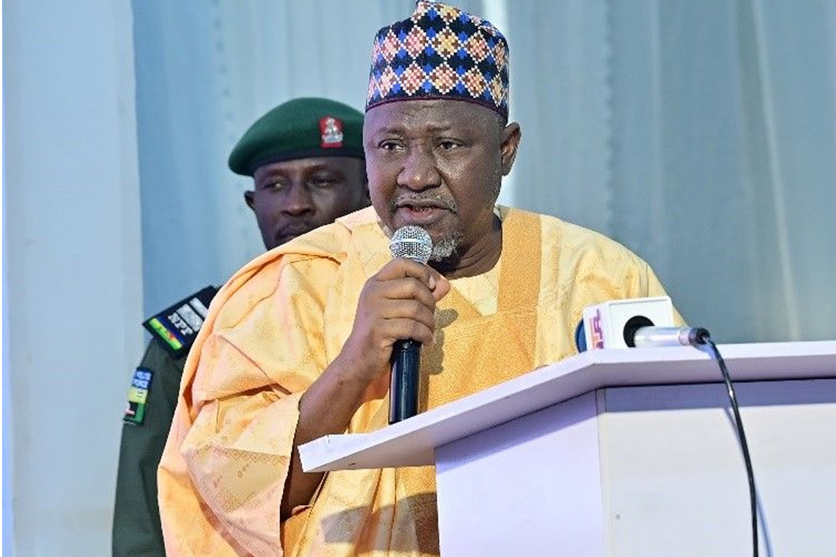 Man in a yellow traditional robe and patterned cap speaks into a handheld microphone at a lectern with press microphones nearby, a security officer in the background
