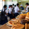 School cafeteria with students in uniform near a counter filled with pizza, burgers, pastries and bottled drinks in the foreground.