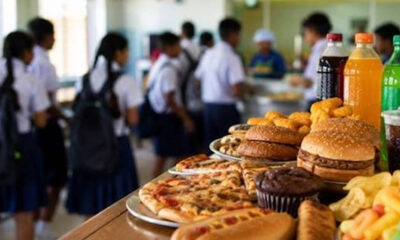 School cafeteria with students in uniform near a counter filled with pizza, burgers, pastries and bottled drinks in the foreground.
