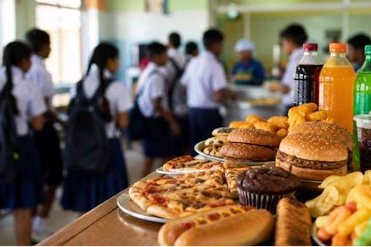 School cafeteria with students in uniform near a counter filled with pizza, burgers, pastries and bottled drinks in the foreground.