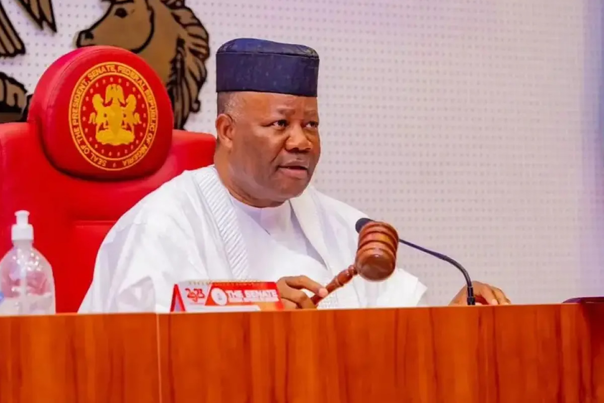 A Nigerian official in white traditional attire sits at a desk with a gavel, in a senate chamber with a red chair behind him.