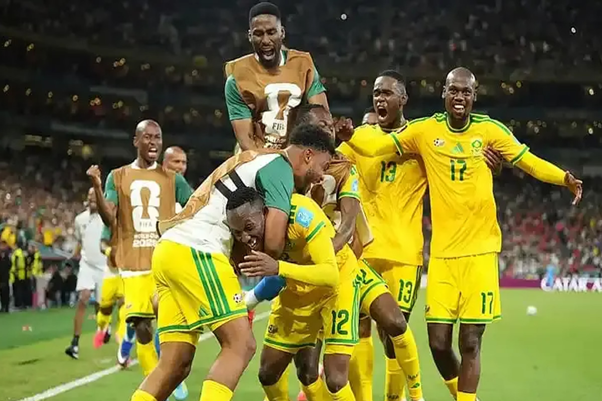 Brazilian soccer players in yellow and green celebrate a goal, hugging and cheering on the pitch as a crowd watches in the stands.
