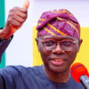 Smiling man in glasses and a purple patterned cap gives a thumbs up beside a red microphone during a speech.