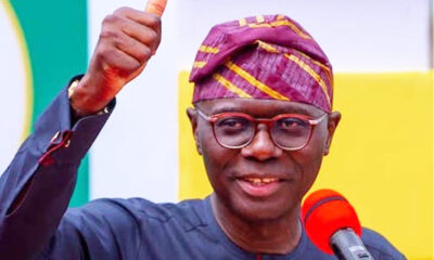 Smiling man in glasses and a purple patterned cap gives a thumbs up beside a red microphone during a speech.