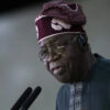 Older man in a dark blue agbada and patterned cap speaks into two microphones, wearing round glasses and a headset.