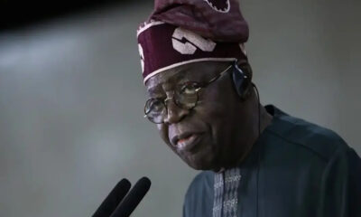 Older man in a dark blue agbada and patterned cap speaks into two microphones, wearing round glasses and a headset.