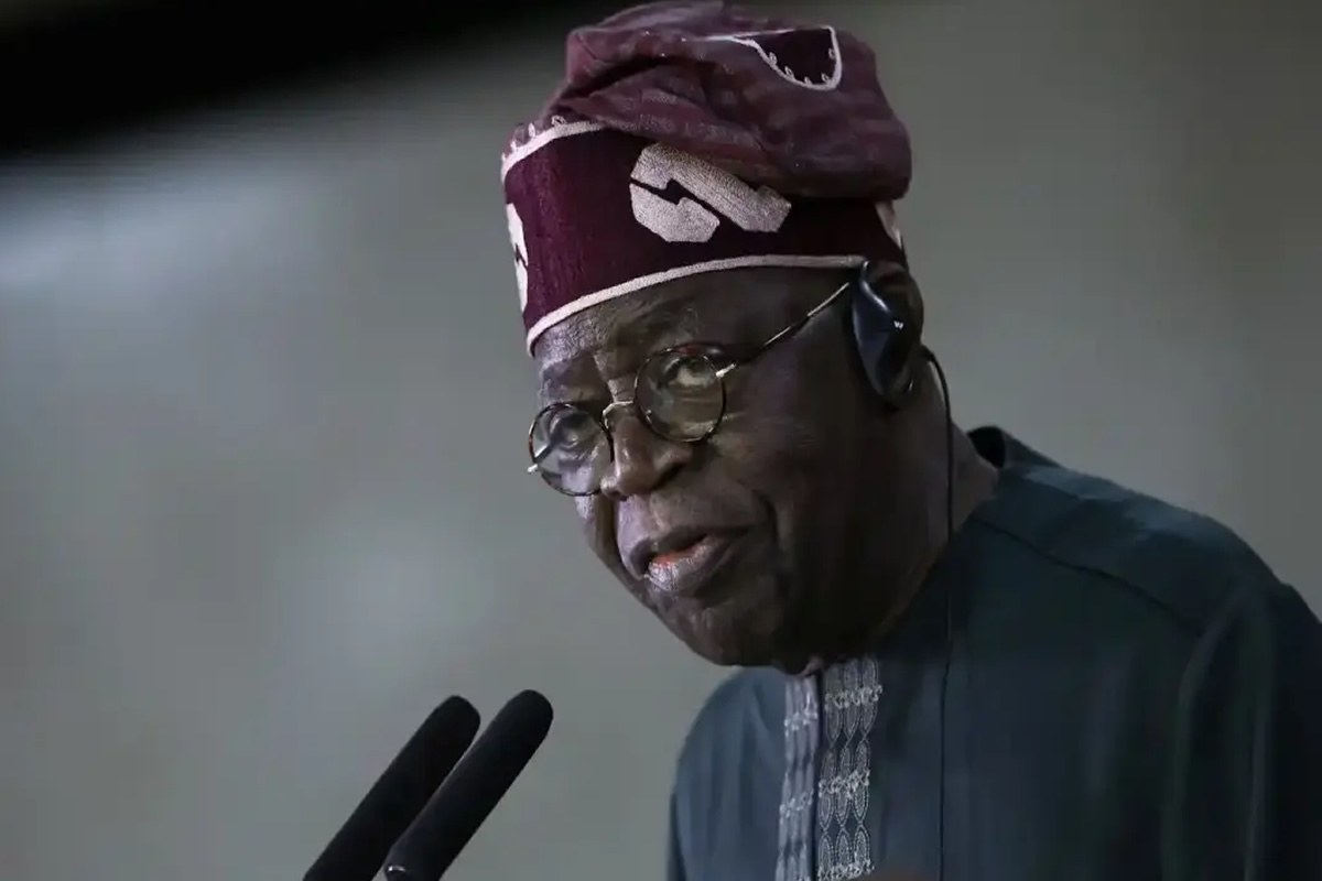 Older man in a dark blue agbada and patterned cap speaks into two microphones, wearing round glasses and a headset.