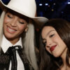 Two smiling women posing closely for a selfie at a party or event; one wears a white cowboy hat and studded jacket, the other has dark hair and red lipstick.