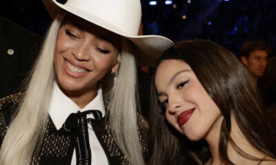 Two smiling women posing closely for a selfie at a party or event; one wears a white cowboy hat and studded jacket, the other has dark hair and red lipstick.