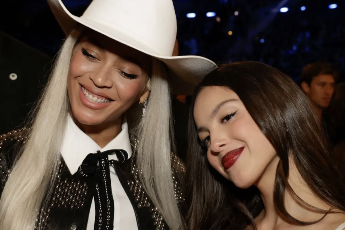 Two smiling women posing closely for a selfie at a party or event; one wears a white cowboy hat and studded jacket, the other has dark hair and red lipstick.
