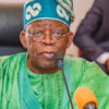 Older man in green traditional outfit and blue cap speaking into a microphone at a formal event.