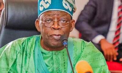 Older man in green traditional outfit and blue cap speaking into a microphone at a formal event.