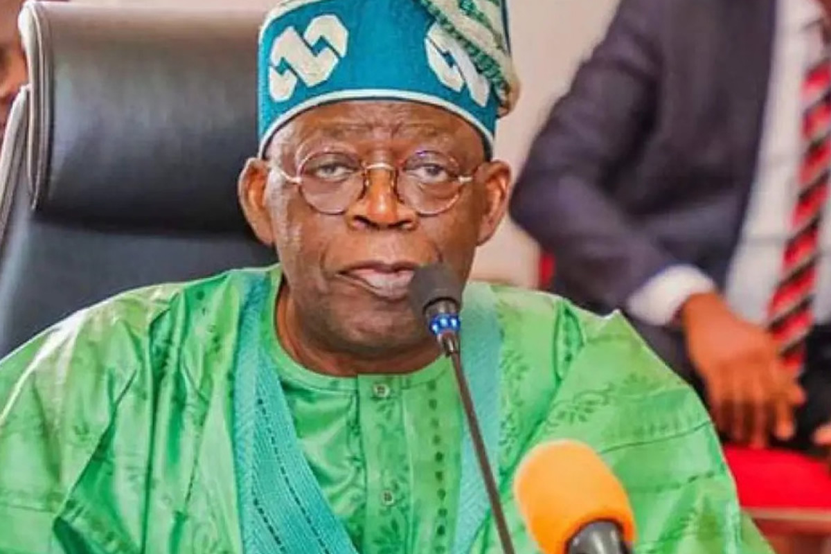 Older man in green traditional outfit and blue cap speaking into a microphone at a formal event.