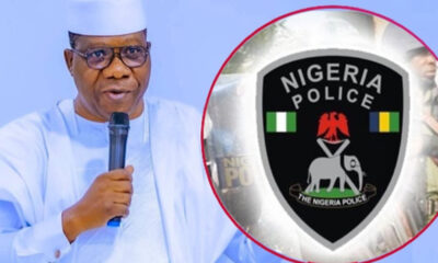 Man in white traditional attire speaks into a microphone with a Nigeria Police emblem overlapping the right side of the image, suggesting a police event or briefing.