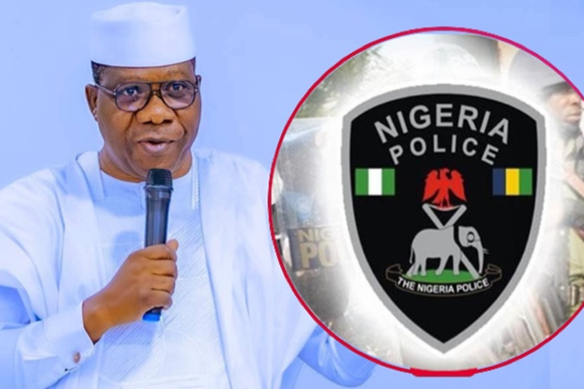 Man in white traditional attire speaks into a microphone with a Nigeria Police emblem overlapping the right side of the image, suggesting a police event or briefing.